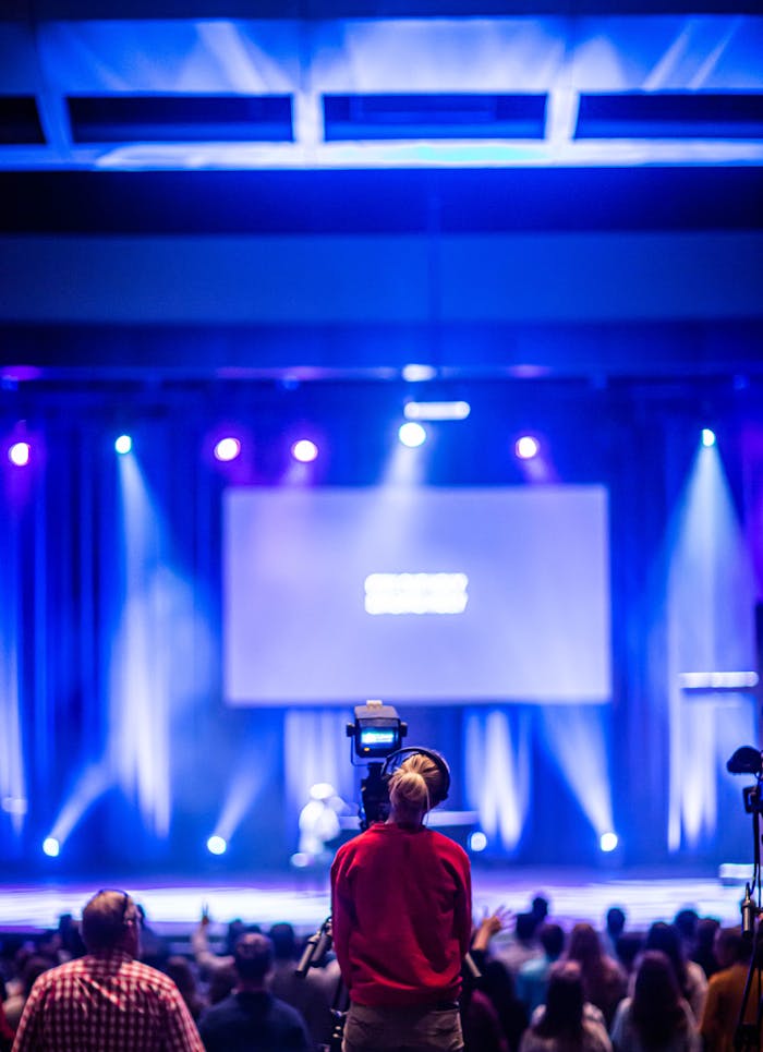 Accueil Cameraman capturing a live event on stage with colorful lights and blurred audience.